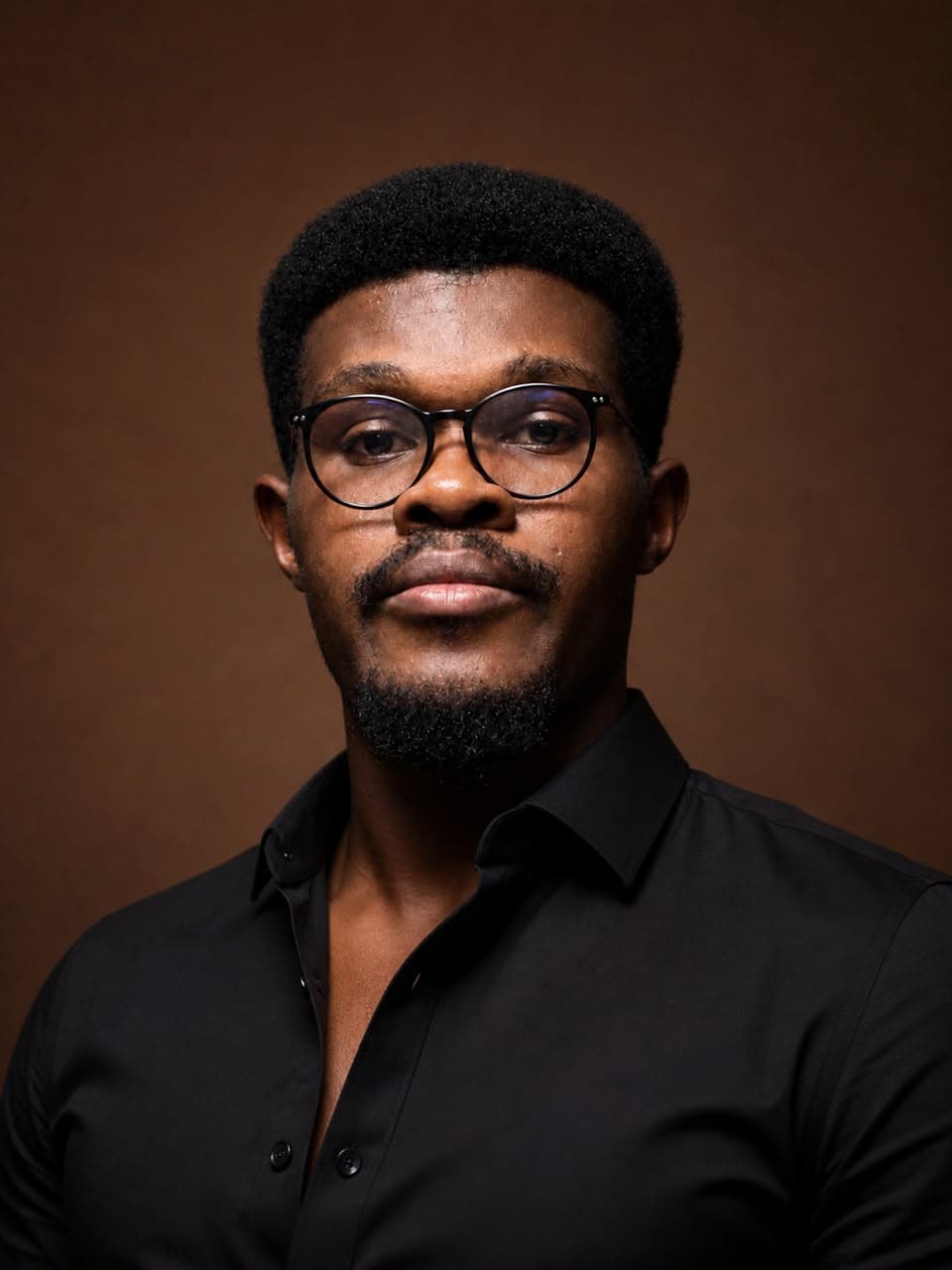 Kingsley Nweke, wearing glasses and a black shirt, photographed against a warm brown backdrop.